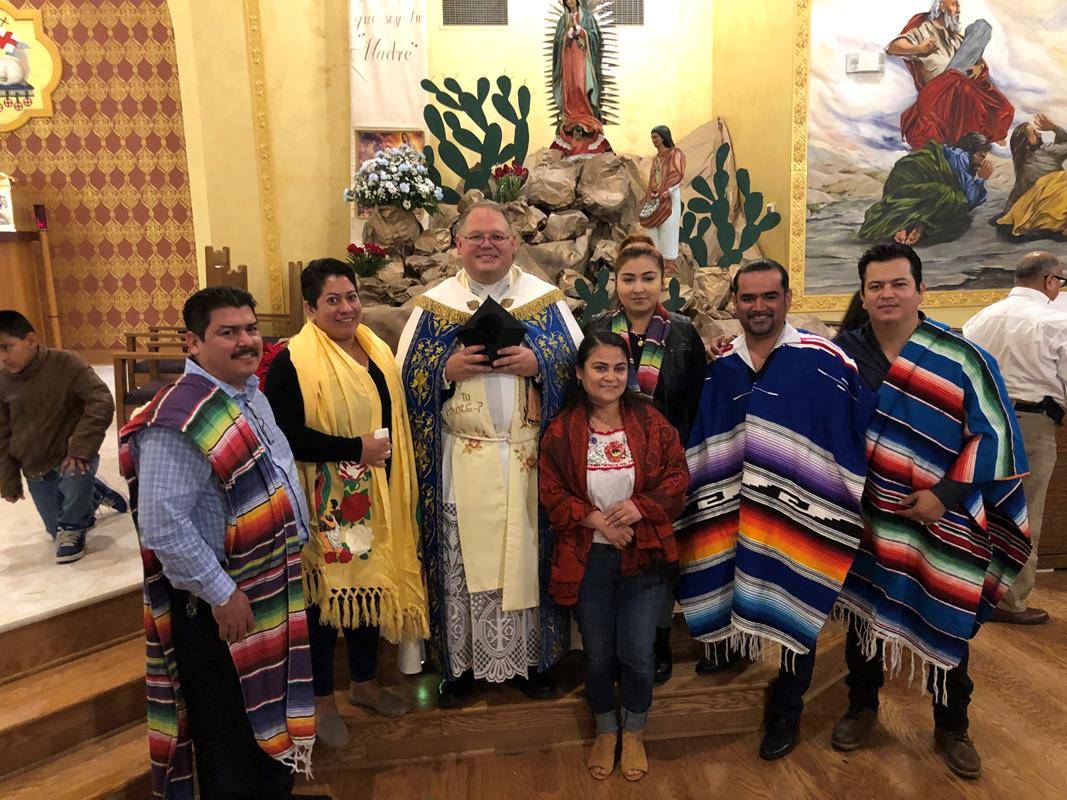 Our Lady of Lourdes parishioners with Father Benjamin Roberts, pastor, at the parish's Feast of Our Lady of Guadalupe Dec. 11. Note the elaborate depiction behind them of Our Lady’s appearance to Juan Diego on the Hill of Tepeyac. (Photo by SueAnn Howell)